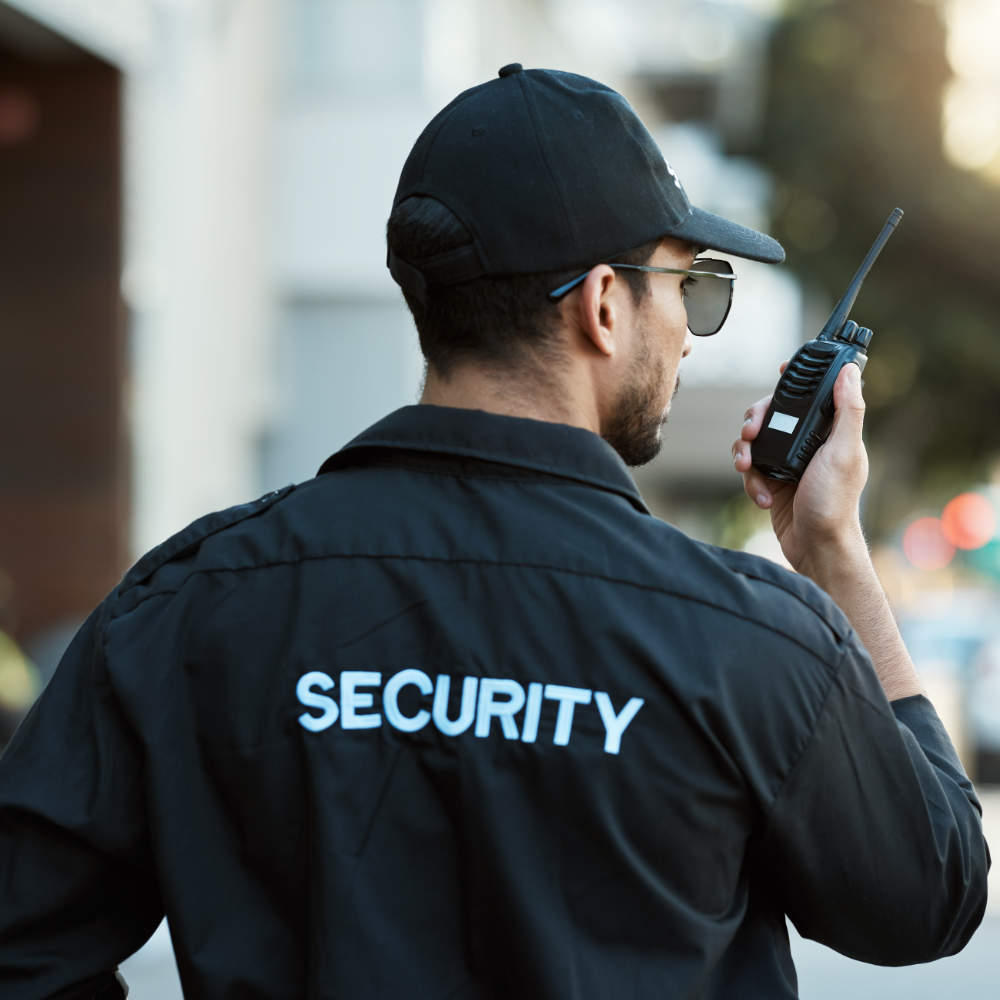 Security guard in a black jacket with 'SECURITY' on the back, using a walkie-talkie.