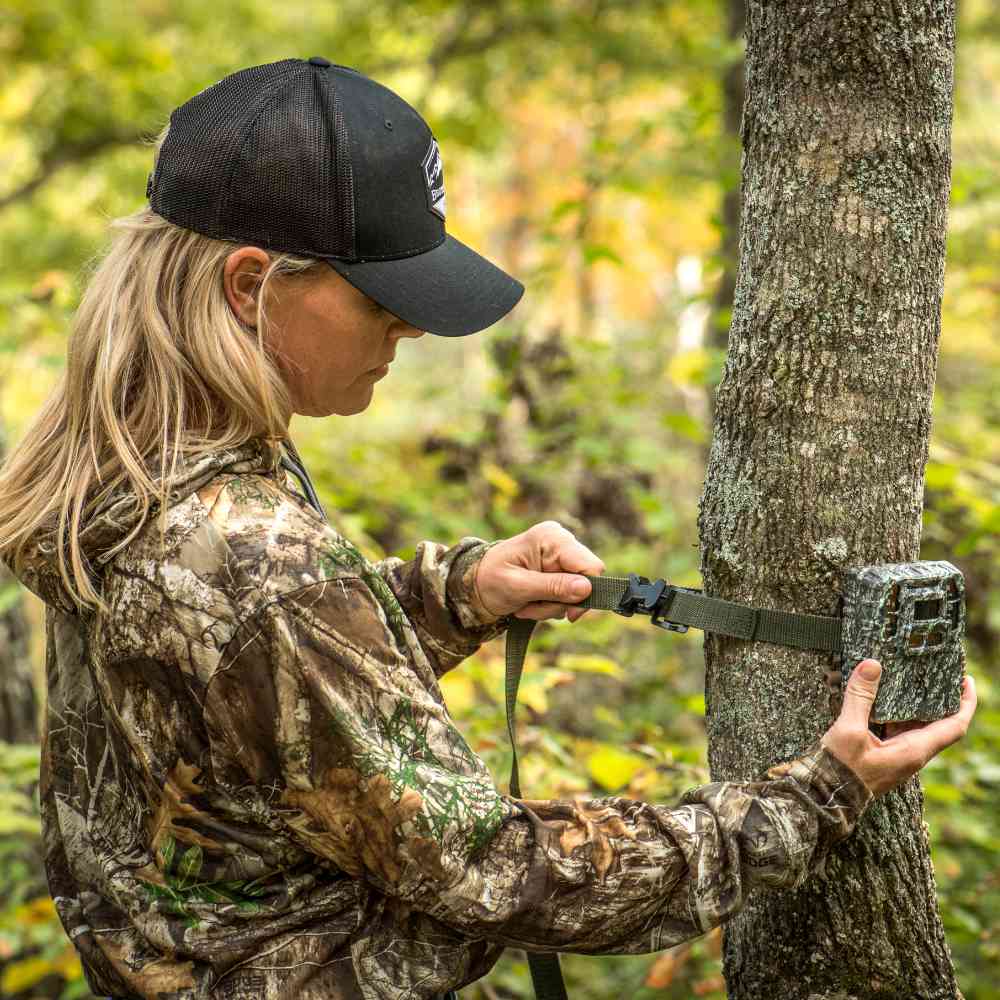 Person in camouflage clothing and black cap using a trail camera in a bush setting