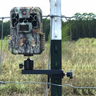 A trail camera mounted on a T-post along a fence in a field.